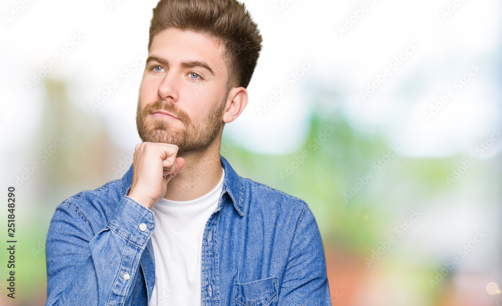 Naklejka premium Young handsome blond man wearing casual denim jacket with hand on chin thinking about question, pensive expression. Smiling with thoughtful face. Doubt concept.