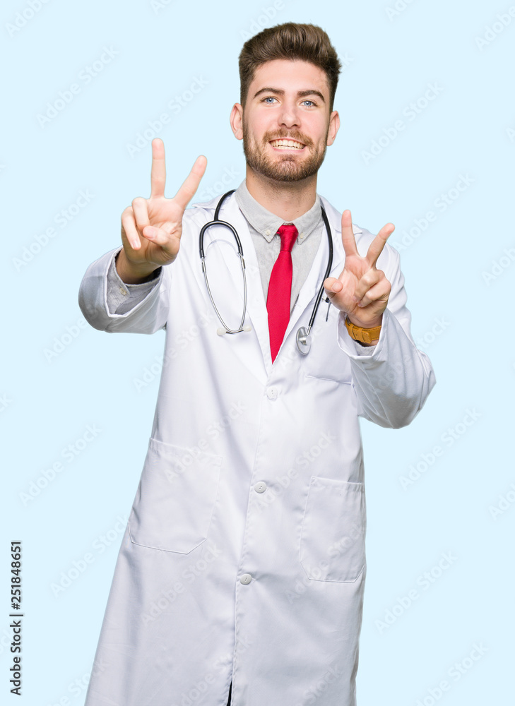 Young handsome doctor man wearing medical coat smiling looking to the camera showing fingers doing victory sign. Number two.