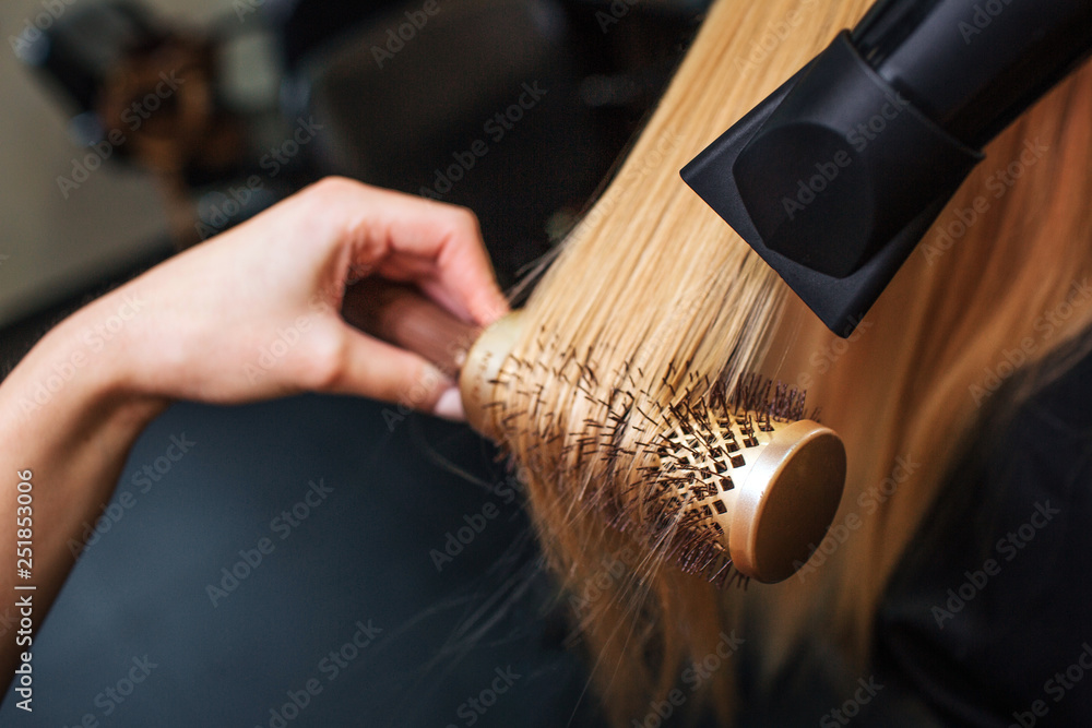 Close-up of hairdressers hand drying blond hair with hair dryer and round brush. Doing hairstyle in beauty salon - obrazy, fototapety, plakaty 