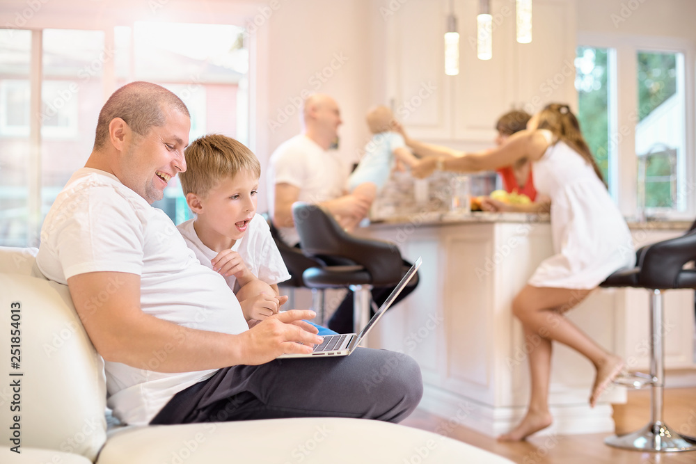 Obraz premium Son with his father sitting on the couch, looking at the laptop, in the kitchen mom friends and baby