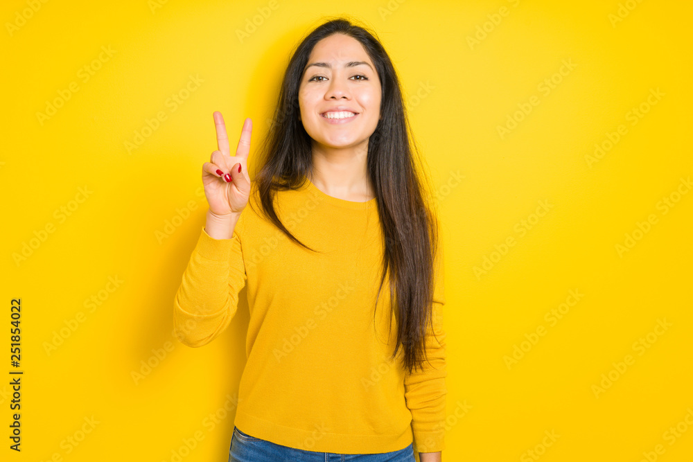 Beautiful brunette woman over yellow isolated background smiling with happy face winking at the camera doing victory sign. Number two. Beautiful brunette woman over yellow isolated background smiling with happy face winking at the camera doing victory sign. Number two.