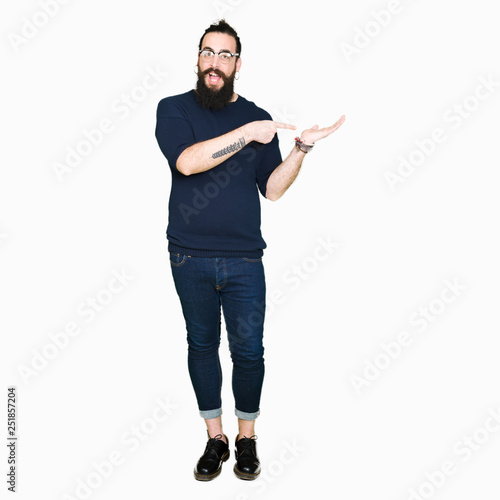 Young hipster man with long hair and beard wearing glasses amazed and smiling to the camera while presenting with hand and pointing with finger.