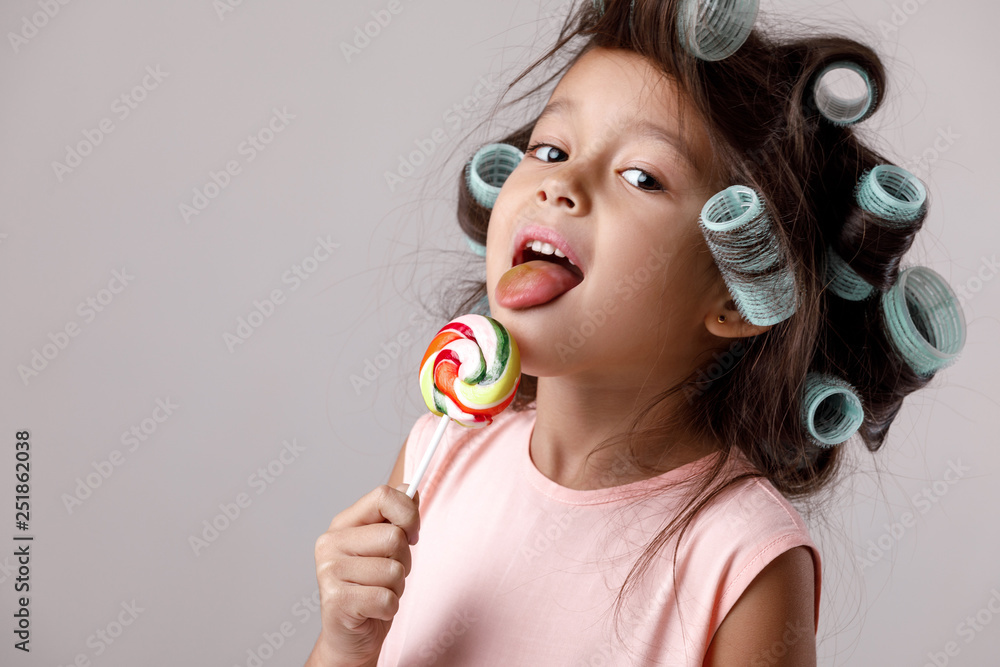 Funny little child girl in pink dress and hair curlers eating lollipop on gray background. Funny little child girl in pink dress and hair curlers eating lollipop on gray background.