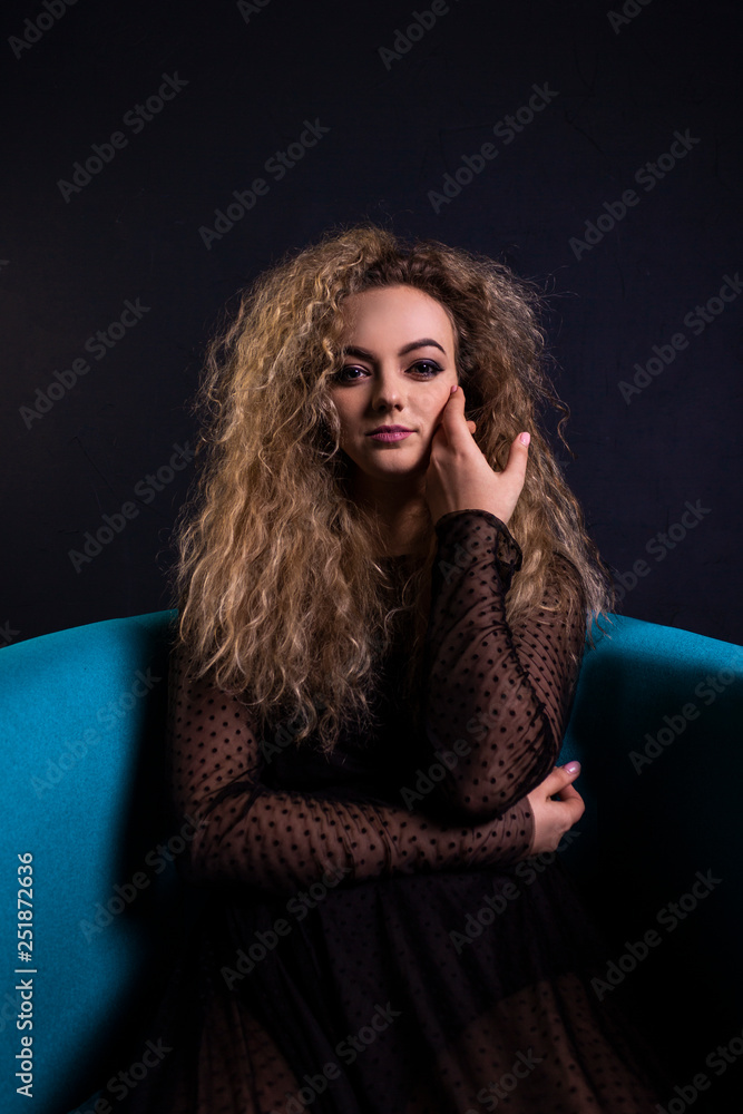 young beautiful sensual girl in a light dark dress with dark curly hair, sitting on the blue chair, in a room with gray walls young beautiful sensual girl in a light dark dress with dark curly hair, sitting on the blue chair, in a room with gray walls