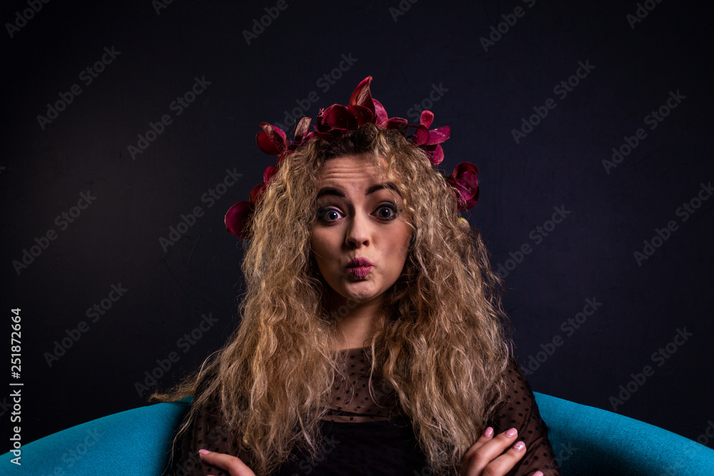 Fototapeta premium young beautiful sensual girl in a light dark dress with dark curly hair, sitting on the blue chair, in a room with gray walls