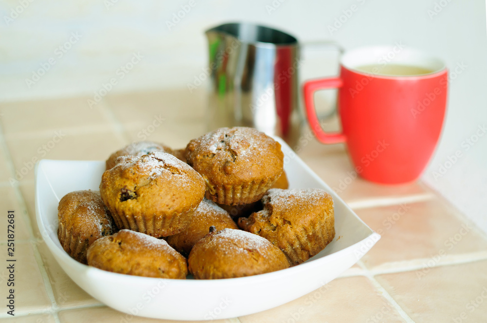 Muffins with powdered sugar on a platter and a milk pitcher with a mug. Muffins with powdered sugar on a platter and a milk pitcher with a mug.