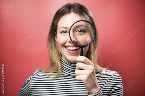 Funny young woman looking through magnifying glass at the camera over pink background.
