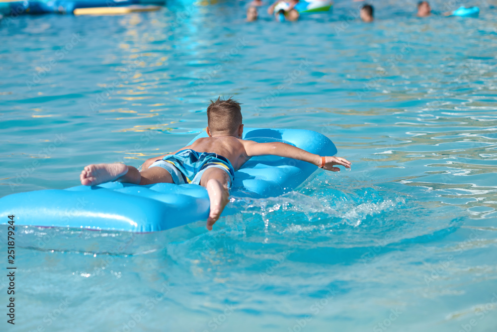 European boy is swimming in the sea on the blue air mattress, making water splashes. He is happy and enjoying his holidays. Back view.