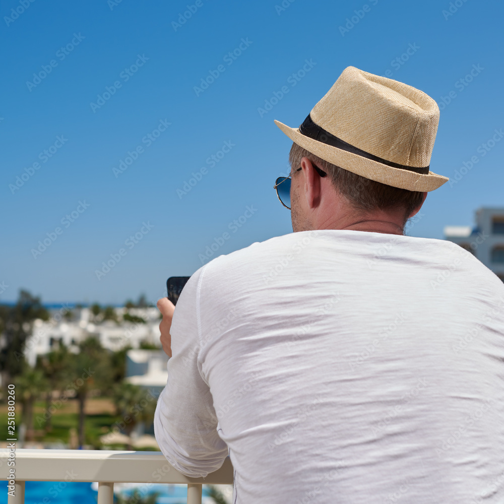 Attractive man in a white t-shirt and sunhat is using his smartphone, while relaxing on the balcony of luxurious hotel.