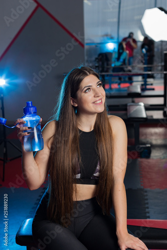 Beautiful sexy fitness model resting after workout on the bench with a bottle of water in hand in the gym.