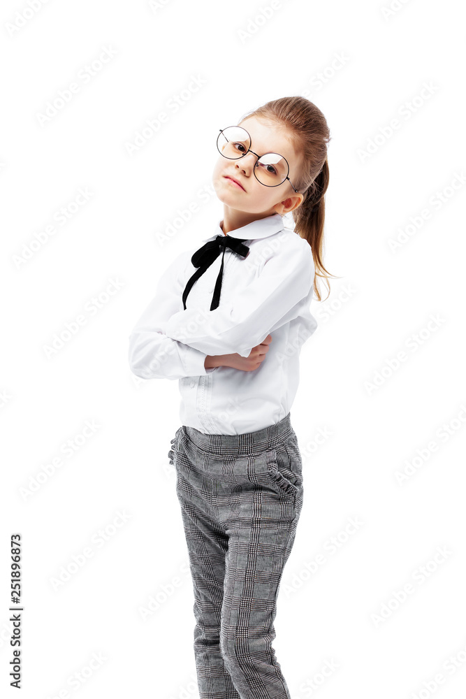 Beautiful little girl dressed like a School girl - in white shirt and gray pants, rounded glasses standing near white wall and posing like model. Beautiful little girl dressed like a School girl - in white shirt and gray pants, rounded glasses standing near white wall and posing like model.