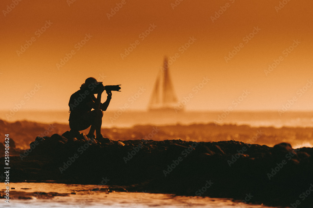 Obraz premium Silhouette of a photographer at the sunset sitting near the sea. Man with photo camera taking pictures at the beach. Sail boat in the background Orange sky and waves. Summer vacation evening landscape