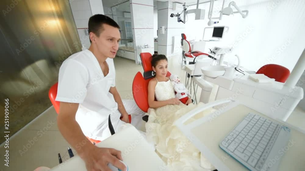 A little girl in a red dental chair at a dentist's reception. A child in a smart dress is talking to a male dentist A little girl in a red dental chair at a dentist's reception. A child in a smart dress is talking to a male dentist