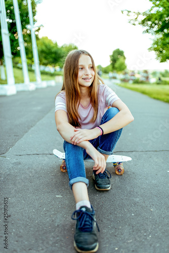 Fototapeta Naklejka Na Ścianę i Meble -  Happy girl teenager 8-12 years old, sitting on a skateboard. In the summer in the city after school lessons resting on the street. Sports schoolgirl in nature looks away.