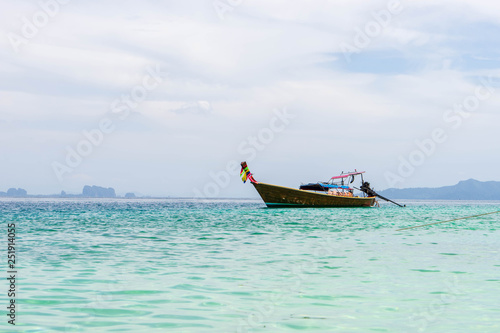 Small wooden tourist boat anchoring on the beach with view of white cloudy sky, island and green clear sea water in Krabi, Southern Thailand.