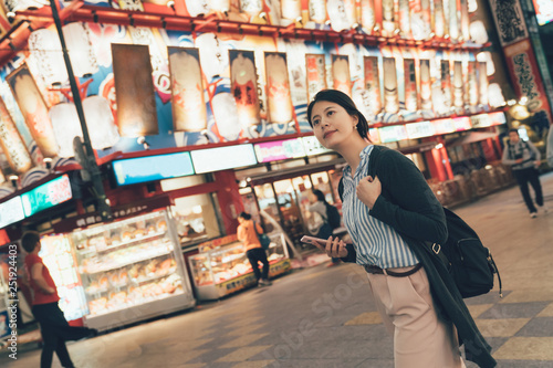 vintage style of beautiful asian woman outdoors at night using mobile phone searching online map walking on urban street. young girl traveler pass by famous seafood restaurant dotonbori osaka japan. photo