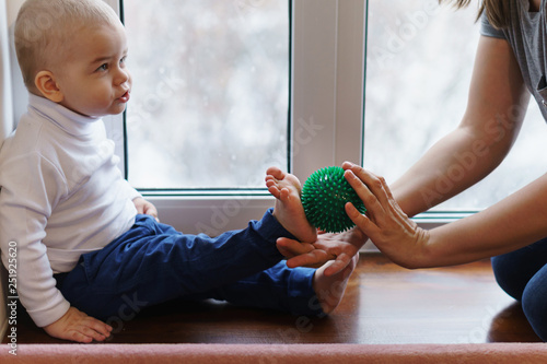 Mother makes baby massage feet. Woman holds massage ball in her hand. Son carefully looks at her. Prevention of flatfoot photo