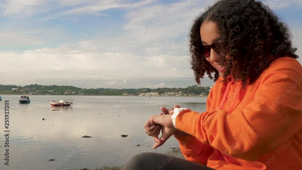 Beautiful mixed race African American girl teenager young woman wearing orange hoodie, sitting on a sea front drinking takeout coffee using her smart watch for social media, communications or texting