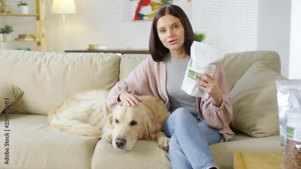 Medium shot of woman sitting on couch, petting dog lying near her, showing packs of dog food and telling information about their quality Medium shot of woman sitting on couch, petting dog lying near her, showing packs of dog food and telling information about their quality