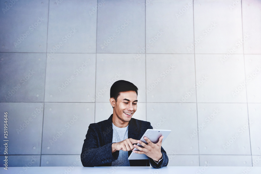 Happy Young Asian Businessman Working on Digital Tablet. Smiling and Sitting at the Desk in Industrial Loft Workplace. Wide Shot Happy Young Asian Businessman Working on Digital Tablet. Smiling and Sitting at the Desk in Industrial Loft Workplace. Wide Shot
