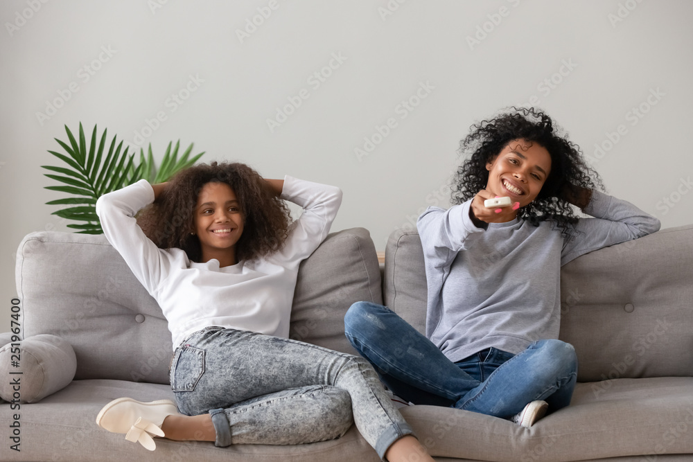 African mother holding remote control resting with daughter on couch