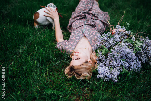 blonde girl in a dress lies on the green grass near the bouquet with lilac and strokes the dog.