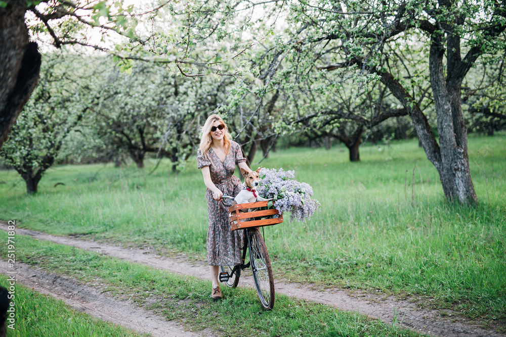 girl blonde in dress and glasses rides on htnny bike with a basket with a dog and flowers in the garden girl blonde in dress and glasses rides on htnny bike with a basket with a dog and flowers in the garden