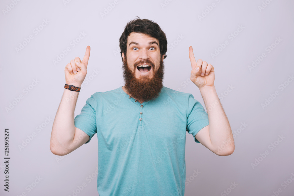 Fototapeta premium Portrait of young surprised handsome bearded man in casual tshirt pointing upside with both hands
