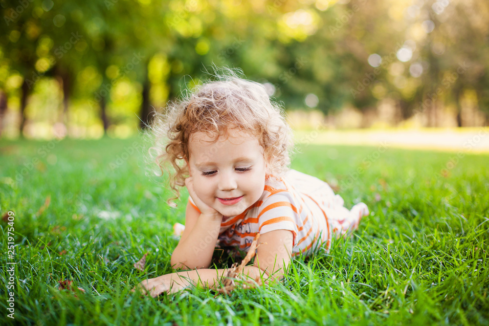 Portrait of adorable little curle girl lying on grass and propping up her face at summer green park Portrait of adorable little curle girl lying on grass and propping up her face at summer green park