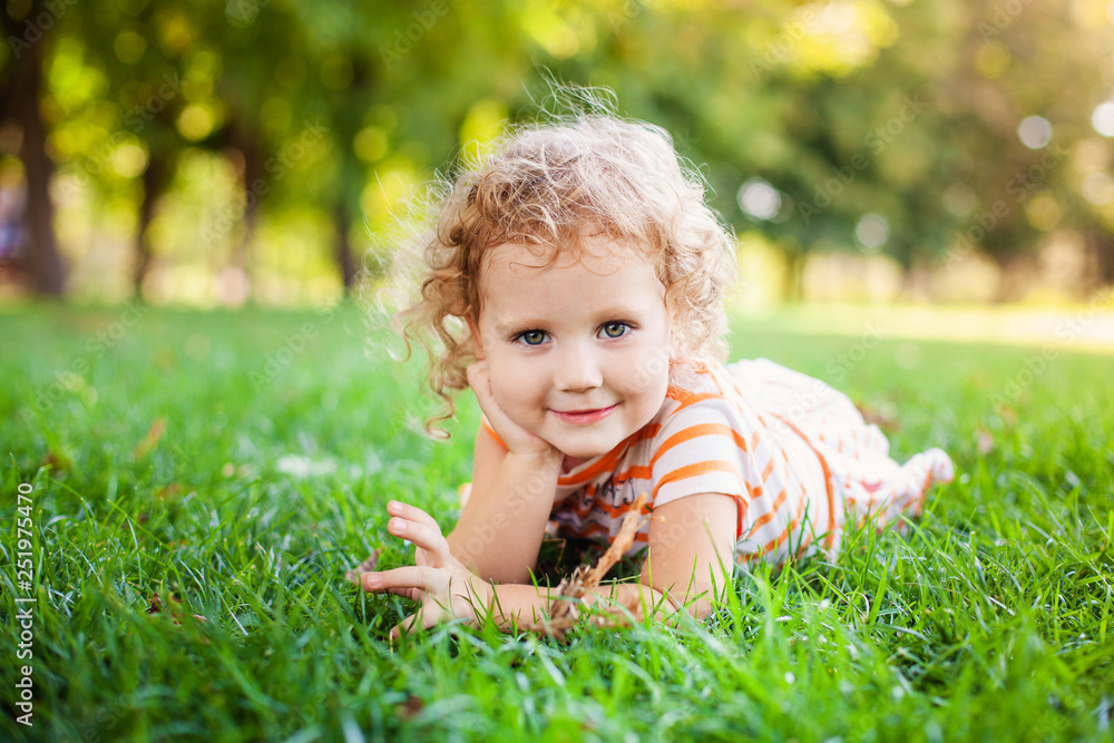 Portrait of adorable little curle girl lying on grass and propping up her face at summer green park Portrait of adorable little curle girl lying on grass and propping up her face at summer green park