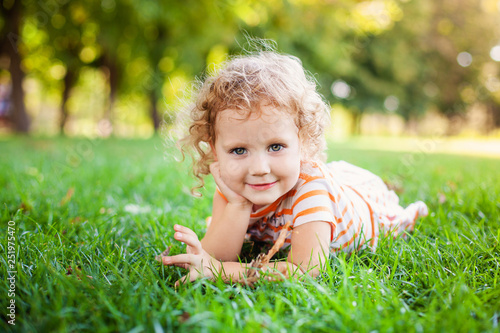 Portrait of adorable little curle girl lying on grass and propping up her face at summer green park