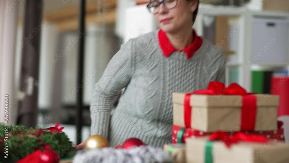 Happy female designer walking in studio, sitting at desk and decorating Christmas wreath with ribbon