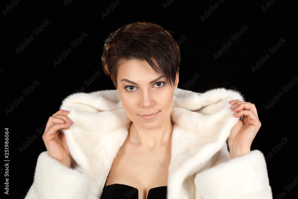 The young beautiful woman in a white fur coat on a black background looks into the camera. The young beautiful woman in a white fur coat on a black background looks into the camera.