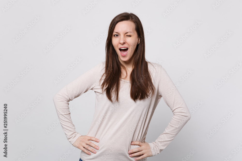 Portrait of funny blinking young woman in light clothes standing with arms akimbo on waist isolated on white wall background in studio. People sincere emotions, lifestyle concept. Mock up copy space. Portrait of funny blinking young woman in light clothes standing with arms akimbo on waist isolated on white wall background in studio. People sincere emotions, lifestyle concept. Mock up copy space.