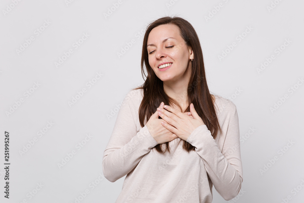 Portrait of pretty young woman in light clothes keeping eyes closed, holding hands crossed on chest isolated on white wall background. People sincere emotions, lifestyle concept. Mock up copy space. Portrait of pretty young woman in light clothes keeping eyes closed, holding hands crossed on chest isolated on white wall background. People sincere emotions, lifestyle concept. Mock up copy space.