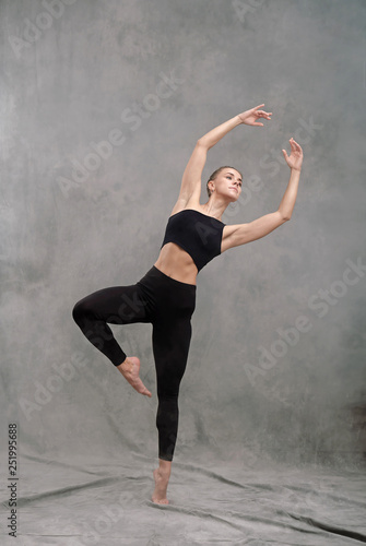 A beautiful graceful slim woman dancer in black clothes performs choreographic figures and movements of modern dance and acrobatics on a gray dark fabric background.