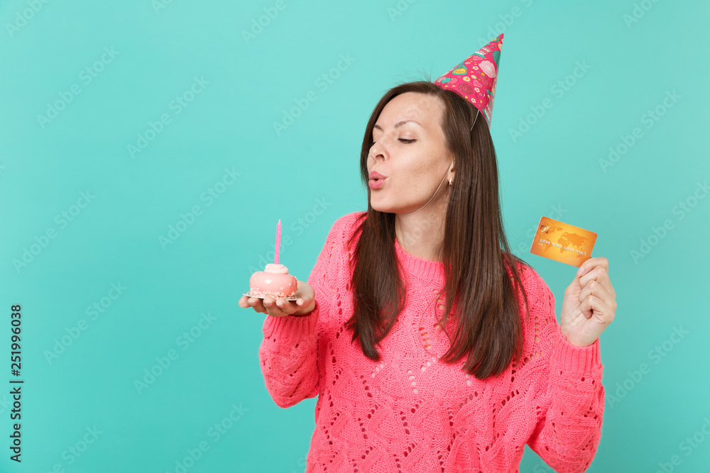 Φωτογραφία Pretty young girl in knitted pink sweater, birthday hat with closed eyes blowing out candle on cake, hold in hand credit card isolated on blue background