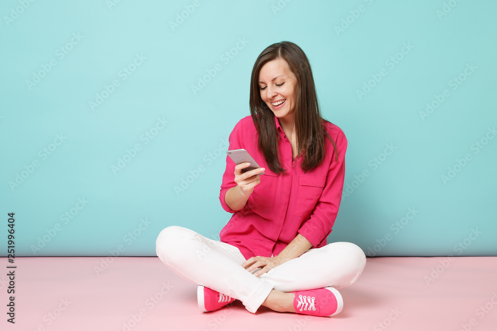 Full length portrait of young fun woman in rose shirt, white pants sit on floor hold cellphone isolated on bright pink blue pastel wall background studio. Fashion lifestyle concept. Mock up copy space Full length portrait of young fun woman in rose shirt, white pants sit on floor hold cellphone isolated on bright pink blue pastel wall background studio. Fashion lifestyle concept. Mock up copy space