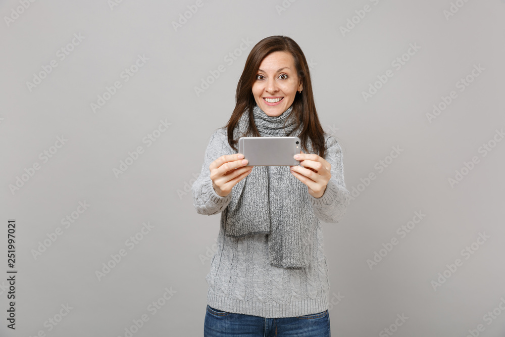 Beautiful young woman in gray sweater, scarf doing taking selfie shot on mobile phone isolated on grey background. Healthy fashion lifestyle, people emotions, cold season concept. Mock up copy space.