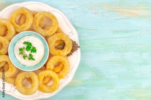 A closeup photo of a plate of calamari rings with a dip and a place for text, shot from the top on a blue background
