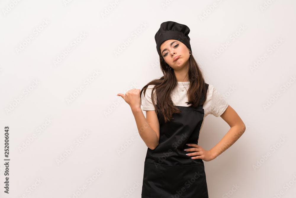 Teenager chef over white wall unhappy and pointing to the side Teenager chef over white wall unhappy and pointing to the side