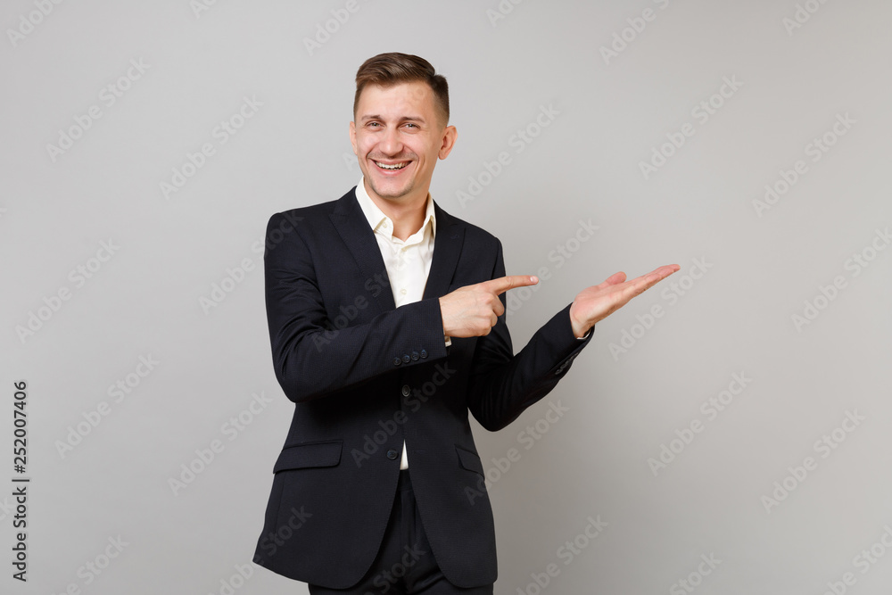 Portrait of joyful young business man in classic black suit pointing index finger hand aside isolated on grey wall background in studio. Achievement career wealth business concept. Mock up copy space. Portrait of joyful young business man in classic black suit pointing index finger hand aside isolated on grey wall background in studio. Achievement career wealth business concept. Mock up copy space.