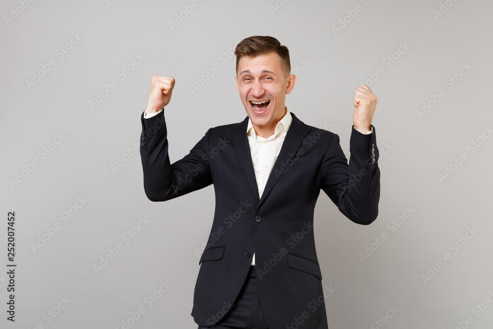 Portrait of laughing young business man in classic black suit, shirt doing winner gesture isolated on grey wall background in studio. Achievement career wealth business concept. Mock up copy space. Portrait of laughing young business man in classic black suit, shirt doing winner gesture isolated on grey wall background in studio. Achievement career wealth business concept. Mock up copy space.