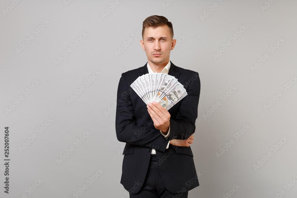 Indifferent young business man in classic black suit holding lots bunch of dollars banknotes, cash money isolated on grey background. Achievement career wealth business concept. Mock up copy space. Indifferent young business man in classic black suit holding lots bunch of dollars banknotes, cash money isolated on grey background. Achievement career wealth business concept. Mock up copy space.