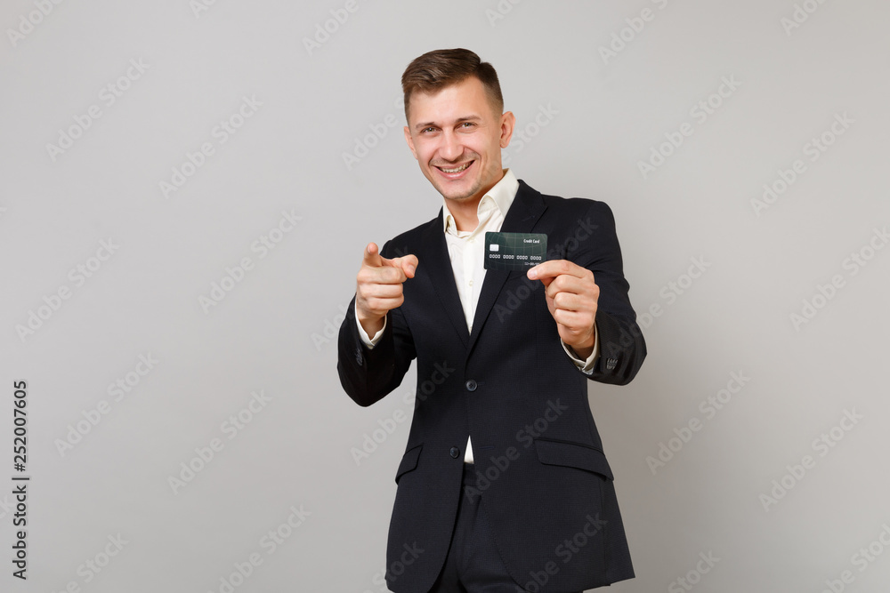 Attractive young business man in classic black suit pointing index finger on camera, hold credit bank card isolated on grey background. Achievement career wealth business concept. Mock up copy space. Attractive young business man in classic black suit pointing index finger on camera, hold credit bank card isolated on grey background. Achievement career wealth business concept. Mock up copy space.