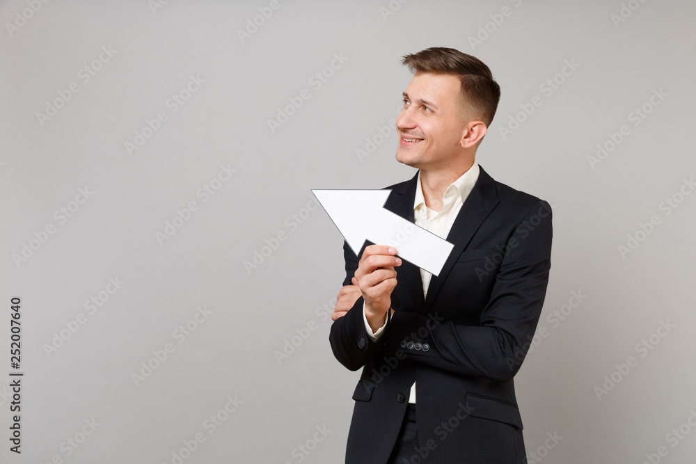 Smiling young business man in classic black suit, shirt looking and showing aside with arrow isolated on grey wall background in studio. Achievement career wealth business concept. Mock up copy space. Smiling young business man in classic black suit, shirt looking and showing aside with arrow isolated on grey wall background in studio. Achievement career wealth business concept. Mock up copy space.