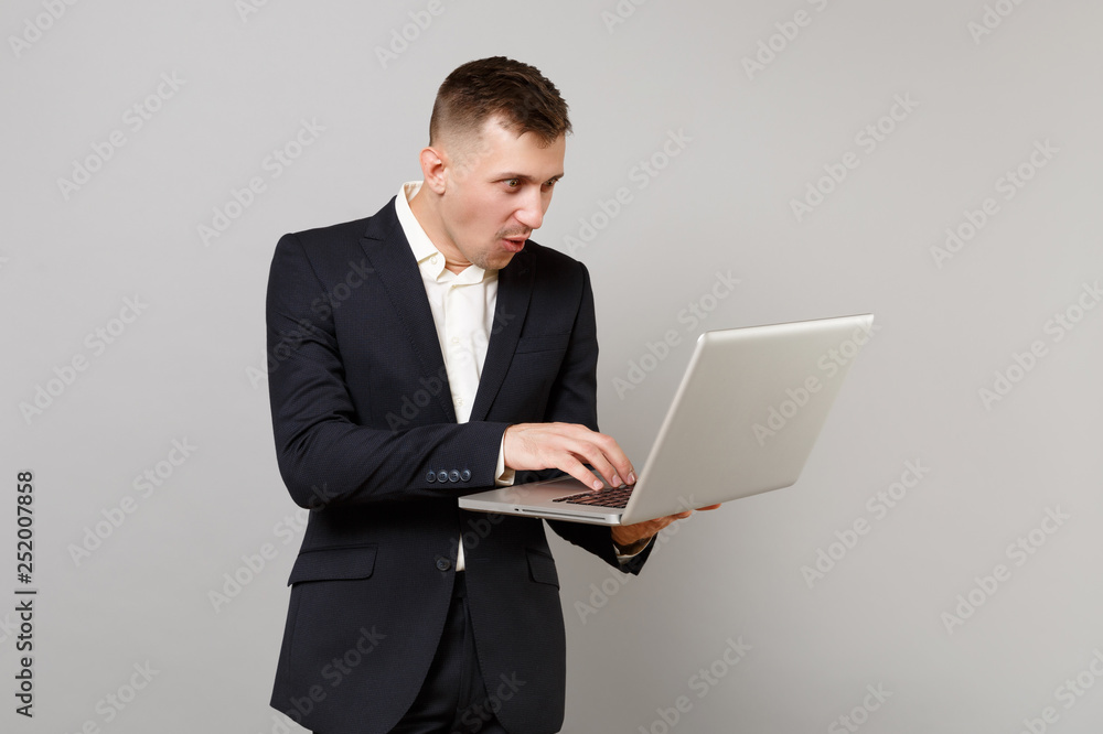Amazed young business man in classic black suit, white shirt working on laptop pc computer isolated on grey wall background in studio. Achievement career wealth business concept. Mock up copy space. Amazed young business man in classic black suit, white shirt working on laptop pc computer isolated on grey wall background in studio. Achievement career wealth business concept. Mock up copy space.