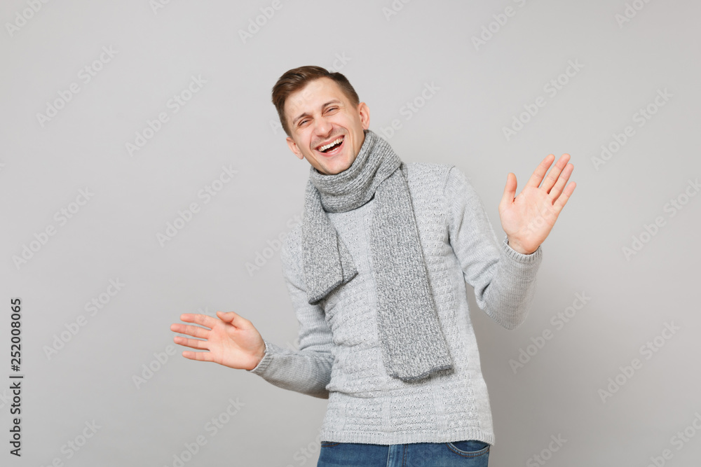 Laughing young man in gray sweater, scarf spreading hands isolated on grey wall background, studio portrait. Healthy fashion lifestyle people sincere emotions, cold season concept. Mock up copy space. Laughing young man in gray sweater, scarf spreading hands isolated on grey wall background, studio portrait. Healthy fashion lifestyle people sincere emotions, cold season concept. Mock up copy space.