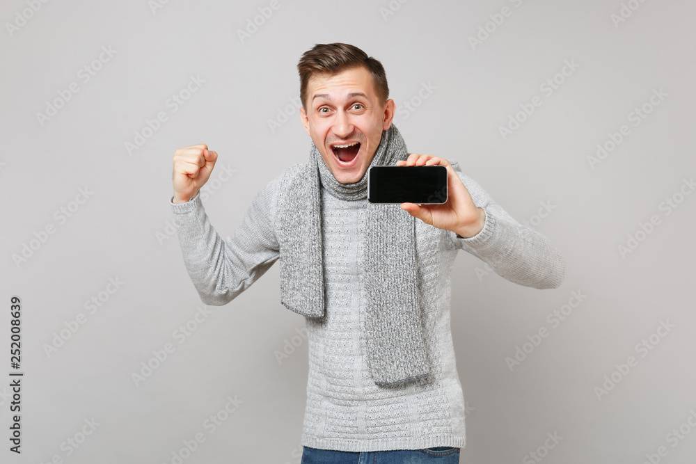 Excited young man in sweater, scarf clenching fist like winner, holding mobile phone with blank empty screen isolated on grey background. Healthy lifestyle people sincere emotions cold season concept.