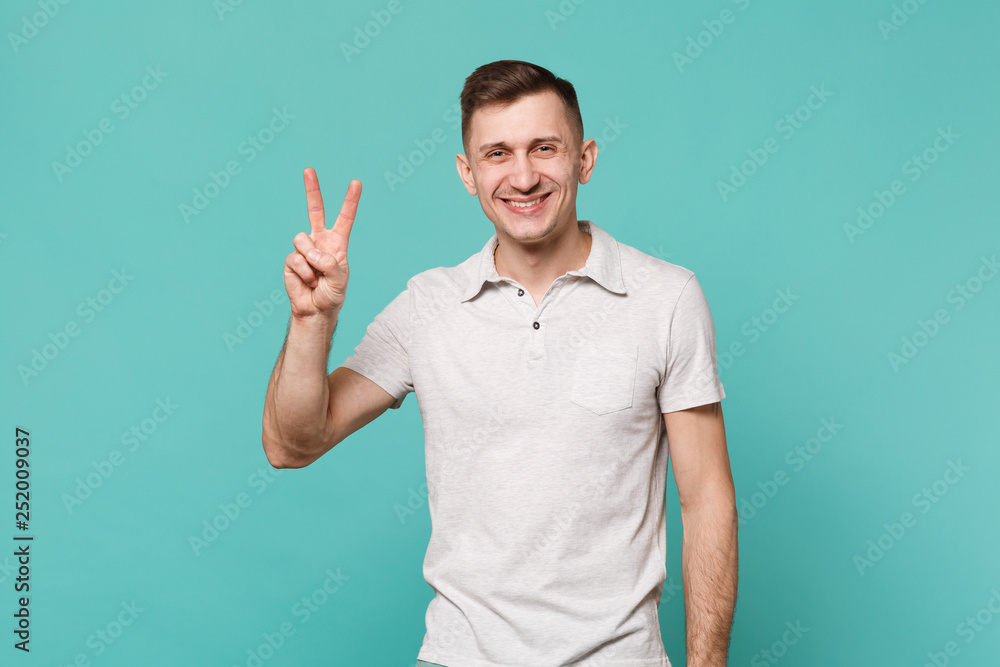 Portrait of attractive smiling young man in casual clothes showing victory sign isolated on blue turquoise wall background in studio. People sincere emotions, lifestyle concept. Mock up copy space. Portrait of attractive smiling young man in casual clothes showing victory sign isolated on blue turquoise wall background in studio. People sincere emotions, lifestyle concept. Mock up copy space.